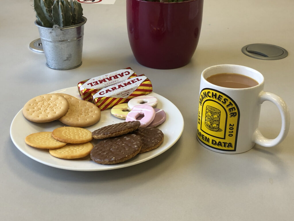 A plate of different biscuits and a mug of tea with Open Data Manchester written on.